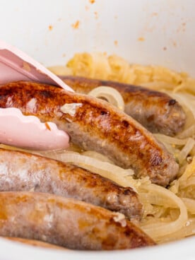 A pair of tongs grabbing a bratwurst from a bed of sauerkraut in a crockpot.