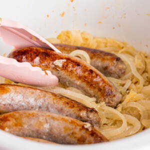 A pair of tongs grabbing a bratwurst from a bed of sauerkraut in a crockpot.