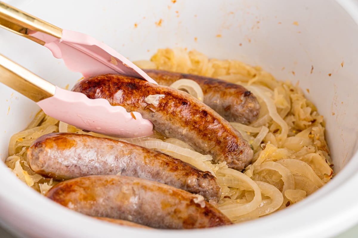 A pair of tongs grabbing a bratwurst from a bed of sauerkraut in a crockpot.