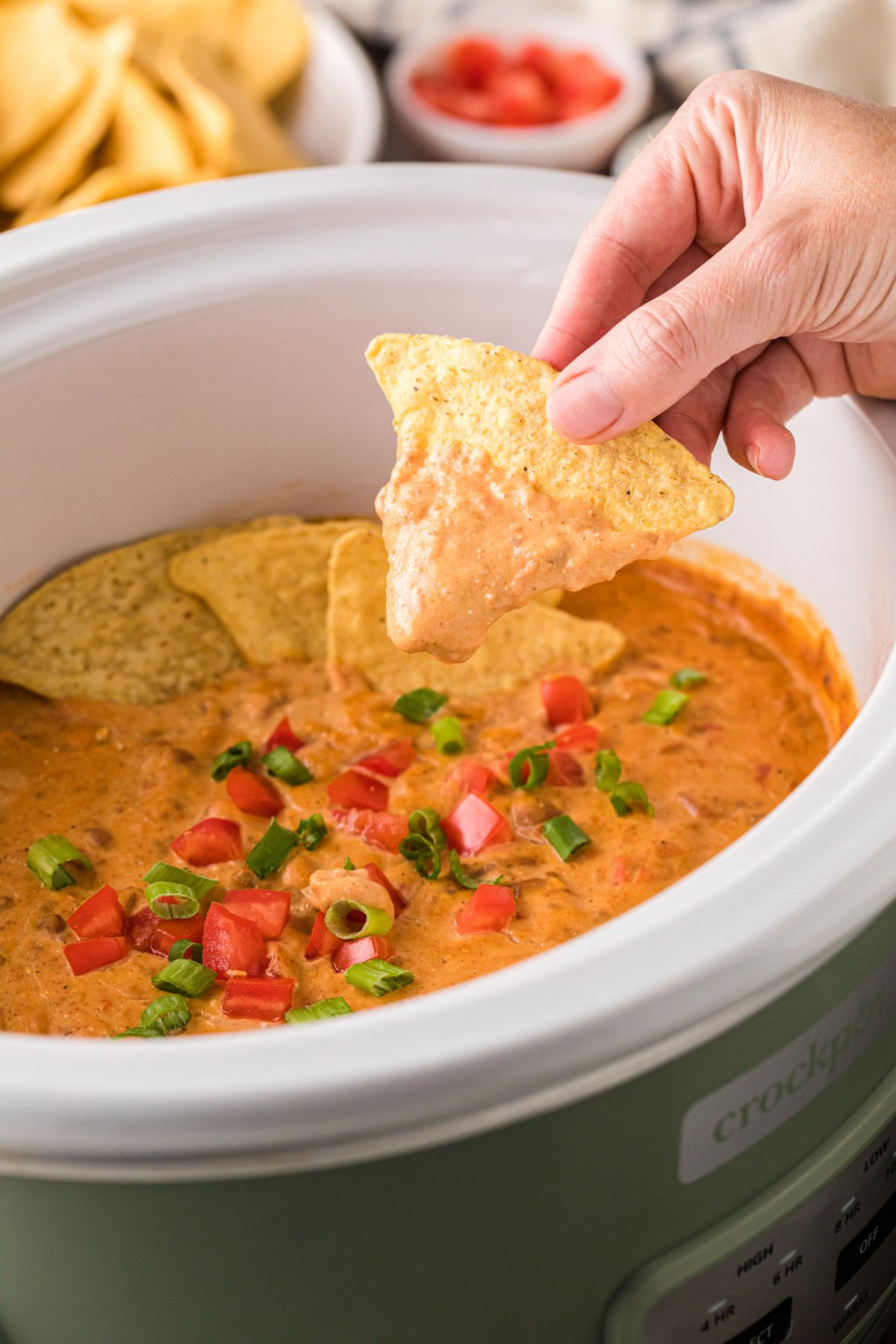 A hand holding a tortilla chip covered in football dip over a crockpot.