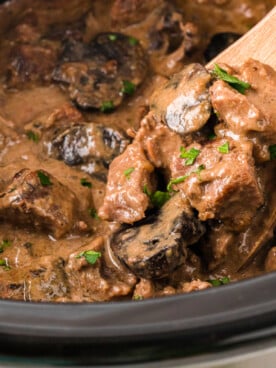 A wooden spoon with a scoop of beef and mushrooms coming out of a crockpot.