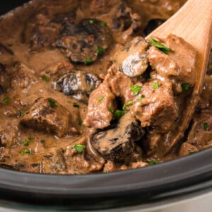 A wooden spoon with a scoop of beef and mushrooms coming out of a crockpot.