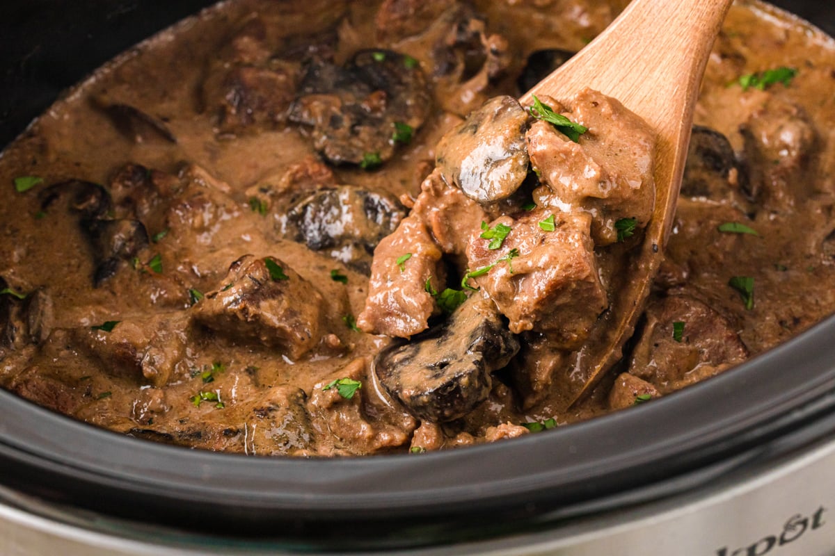 A wooden spoon with a scoop of beef and mushrooms coming out of a crockpot.