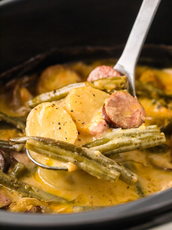 A large spoon holding up a scoop of potatoes, green beans, and kielbasa over a crockpot.