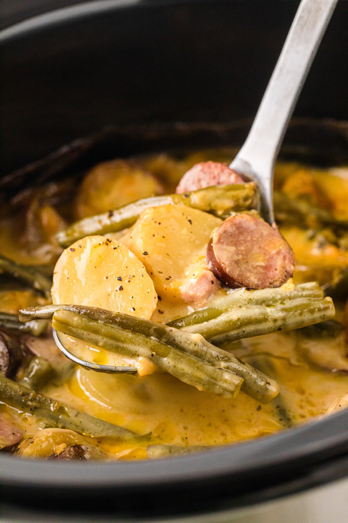 A large spoon holding up a scoop of potatoes, green beans, and kielbasa over a crockpot.