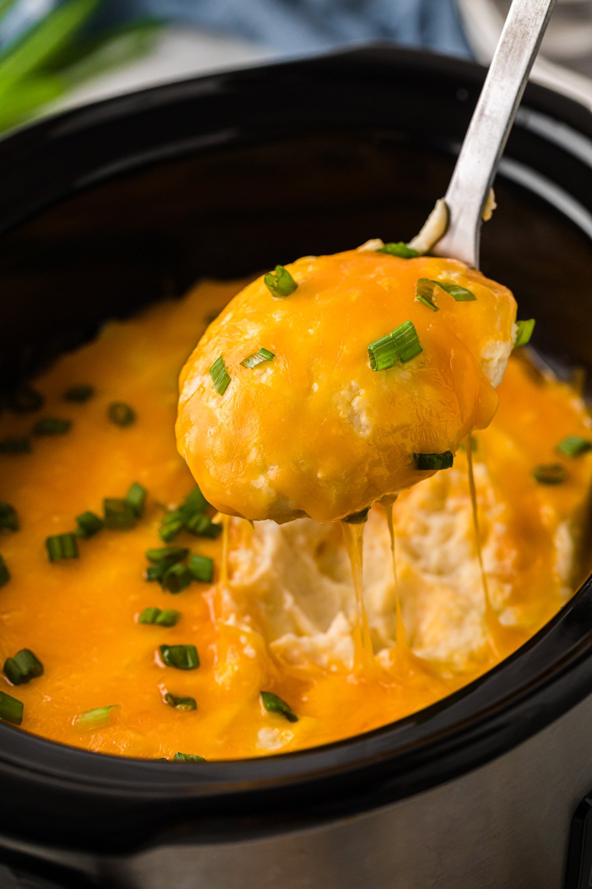 A spoon pulling cheesy mashed potatoes out of a crockpot.
