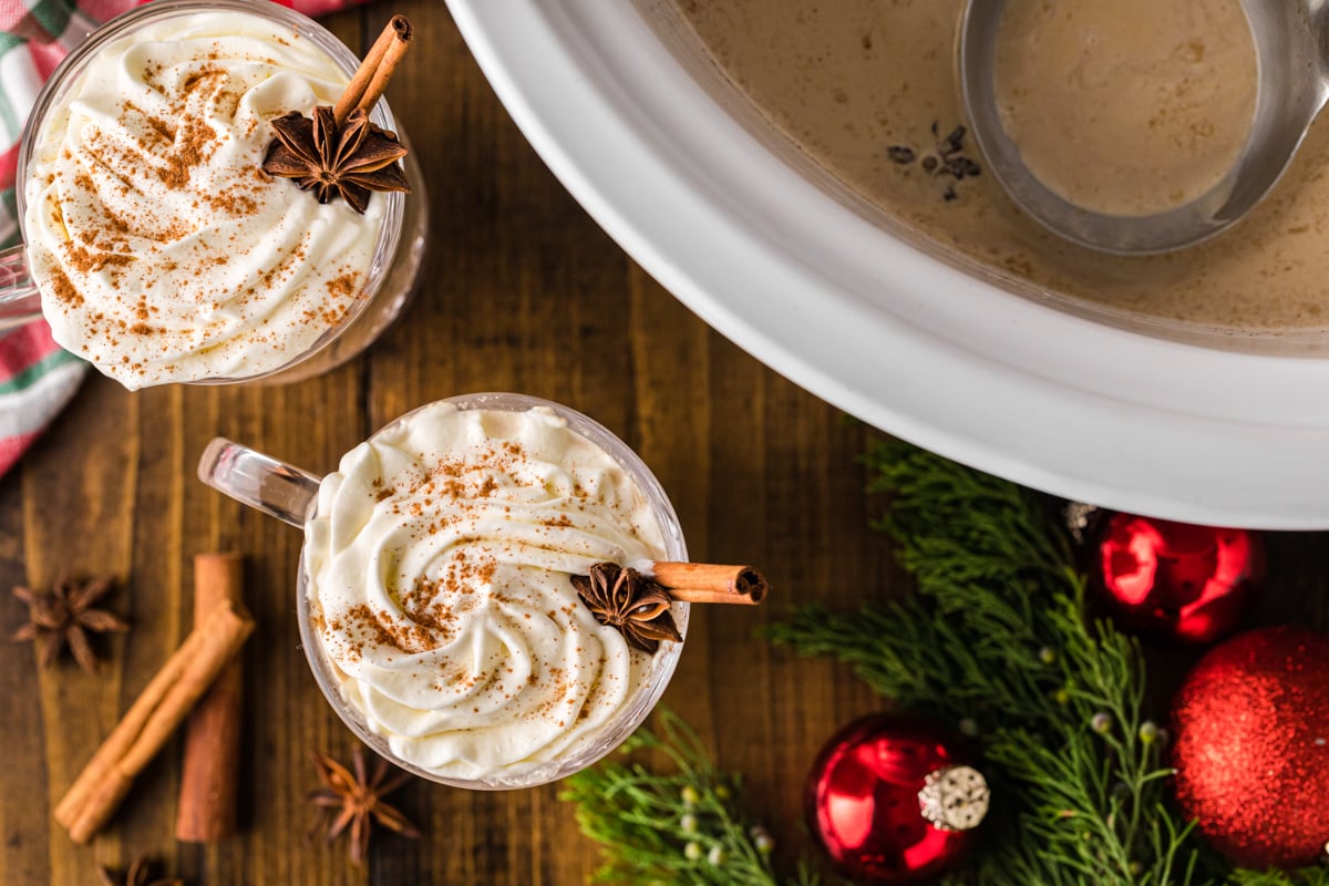 Two mugs of coffee next to a crockpot with Christmas decorations on the table.