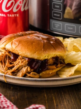 A sandwich with shredded coca cola chicken on a plate with potato chips.