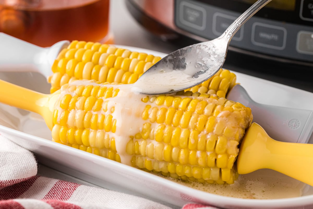 A spoon drizzling coconut milk, honey, and melted butter mixture onto an ear of corn.