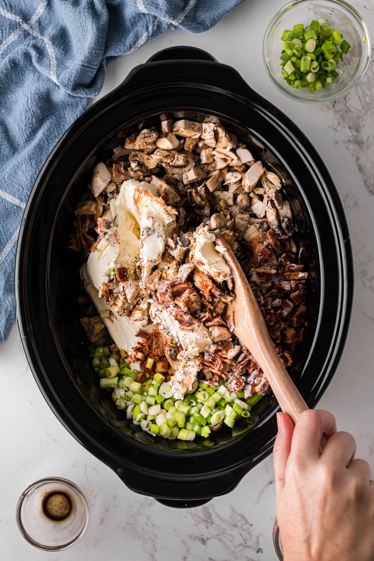 A hand holding a wooden spoon mixing ingredients together in a slow cooker.