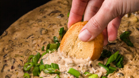 A hand dipping toasted bread into a crockpot of mushroom dip.