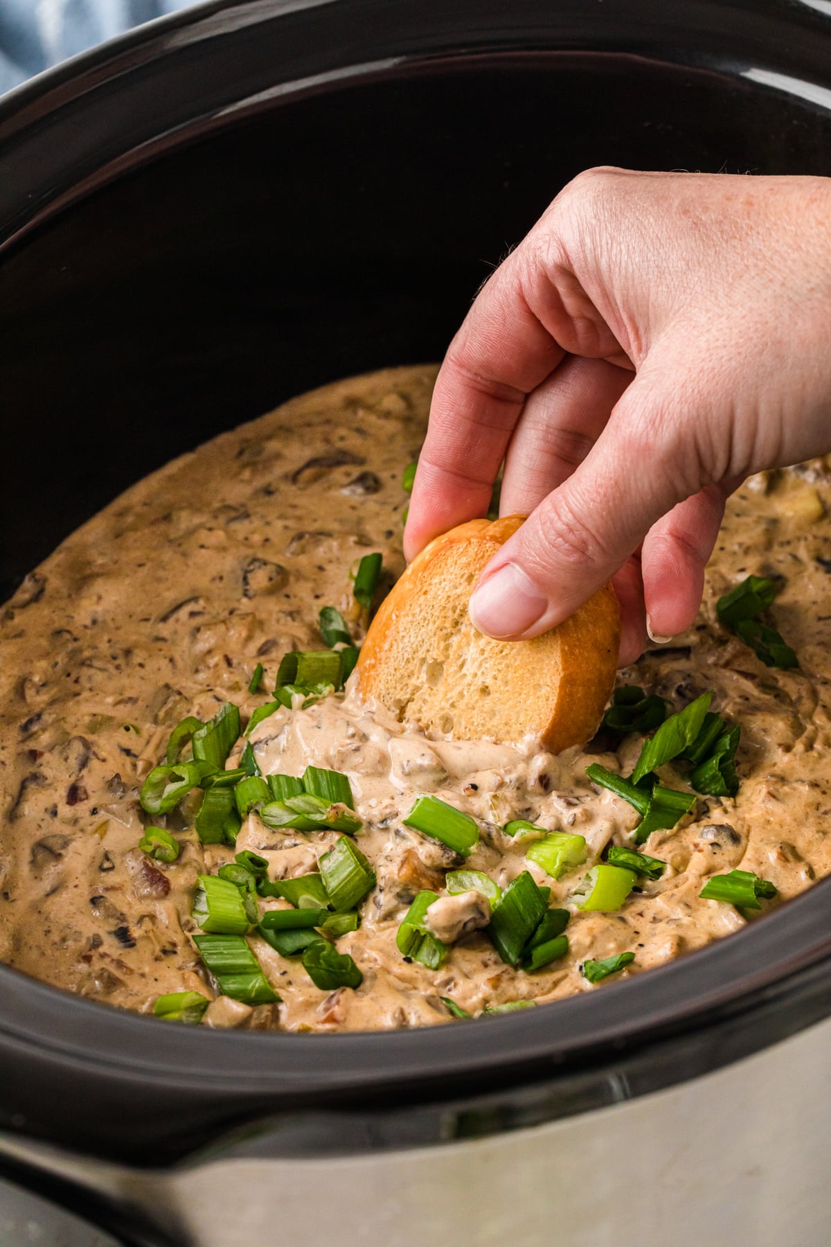 A hand dipping toasted bread into a crockpot of mushroom dip.