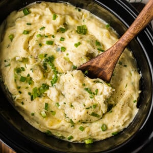 Sour cream and onion mashed potatoes in a slow cooker with a wooden spoon in it.