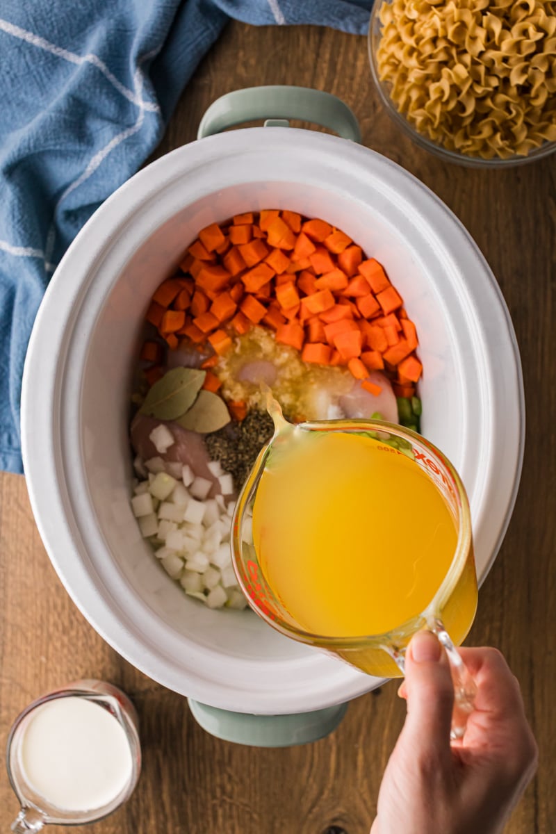 Pouring chicken broth over ingredients in a slow cooker.