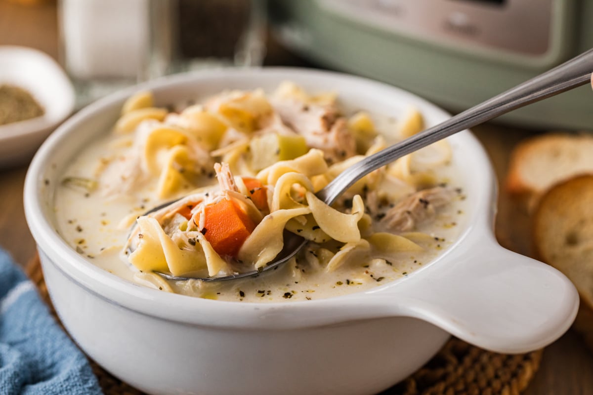 A spoonful of soup coming out of a white bowl in front of a crockpot.