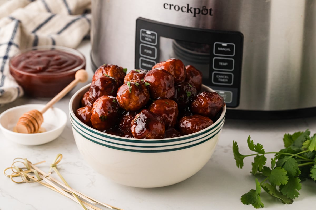 Meatballs in a white bowl with blue lines in front of a crockpot.
