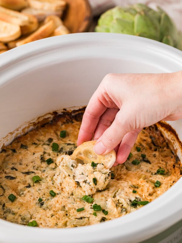 Dipping a toasted baguette piece into crab spinach artichoke dip in a slow cooker.
