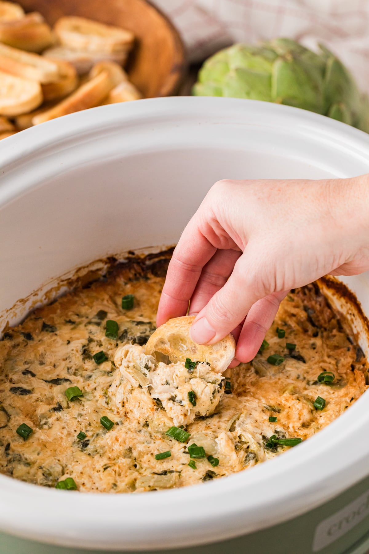 Dipping a toasted baguette piece into crab spinach artichoke dip in a slow cooker.