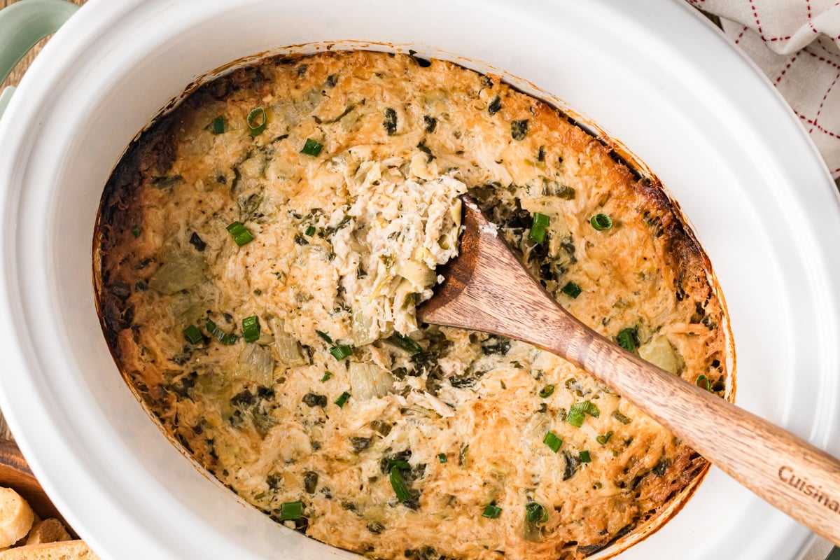A crockpot filled with crab spinach artichoke dip with a wooden spoon in it.