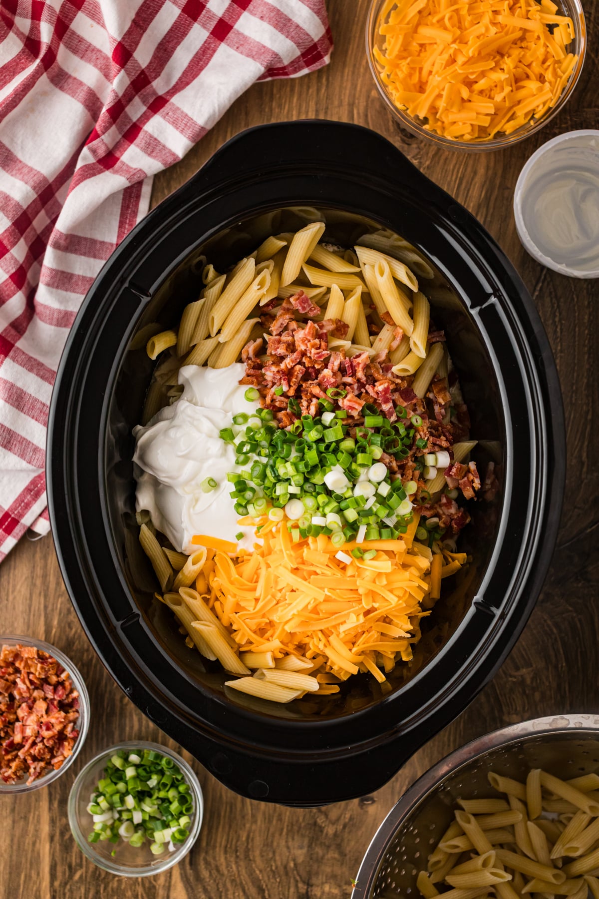 Pasta, cheese, bacon, sour cream, and green onions added to cooked chicken in a crockpot.
