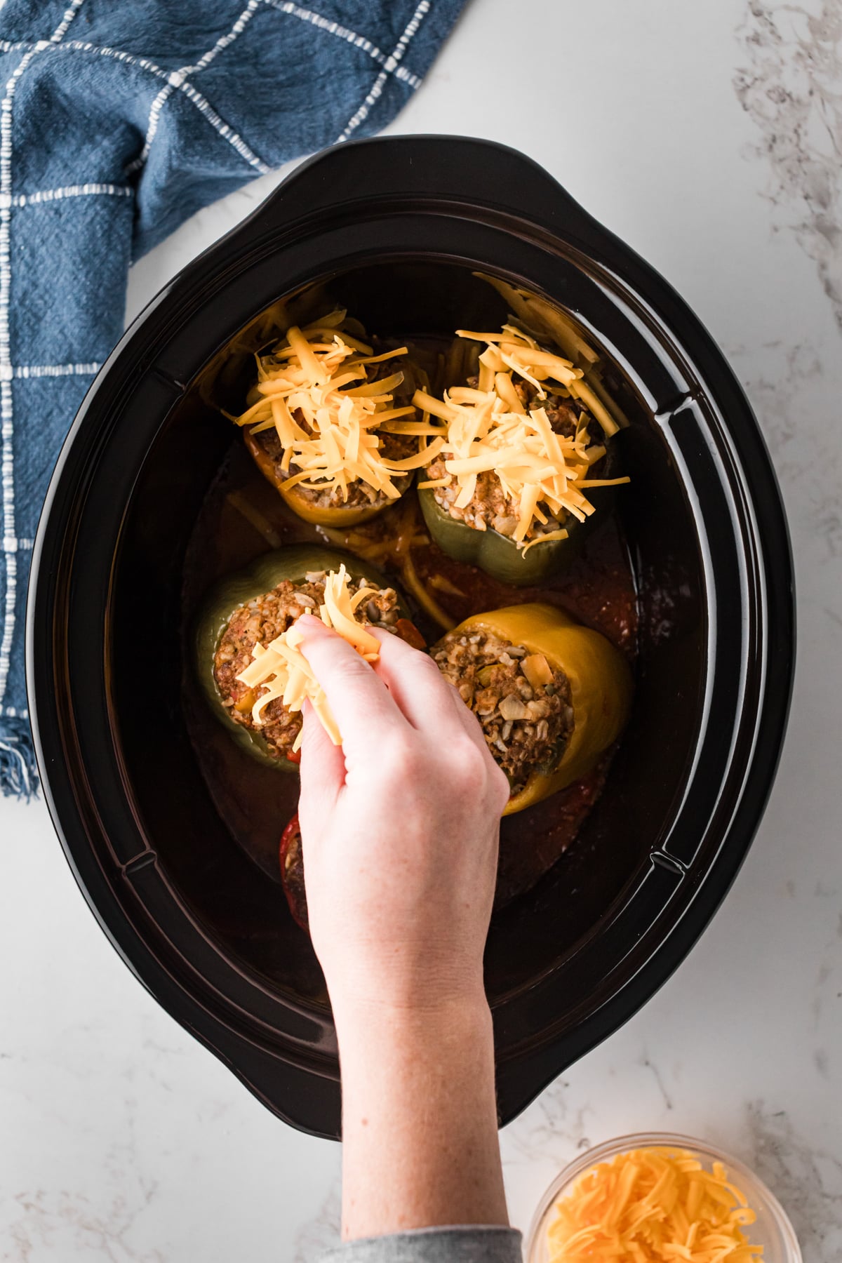 A hand sprinkling cheese onto stuffed bell peppers in a slow cooker.