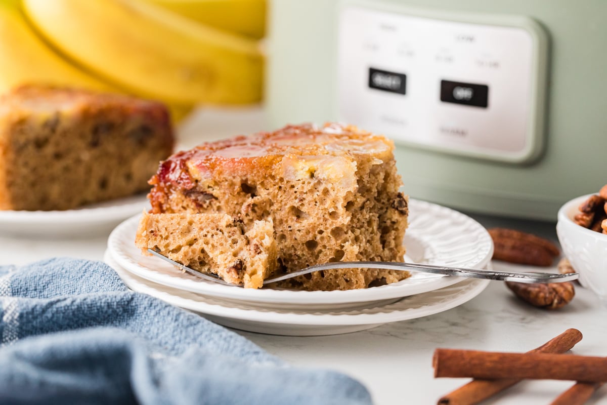 A slice of banana upside down cake on a stack of white plates in front of a crockpot.