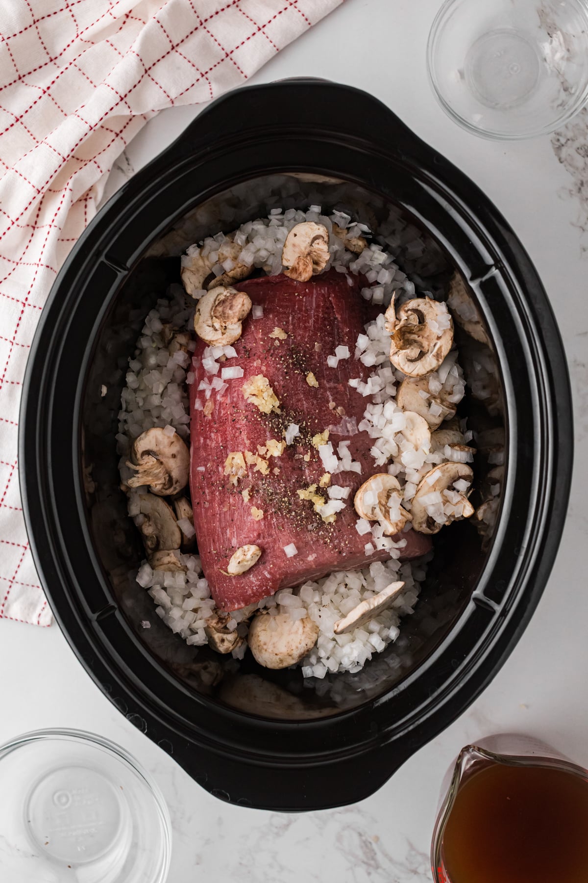 Uncooked seasoned beef roast, garlic, onion, and mushrooms in a crockpot.