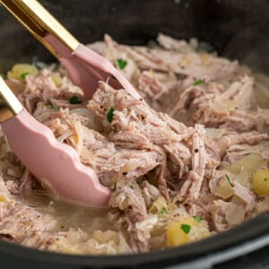 Tongs grabbing some pork and sauerkraut from a slow cooker.