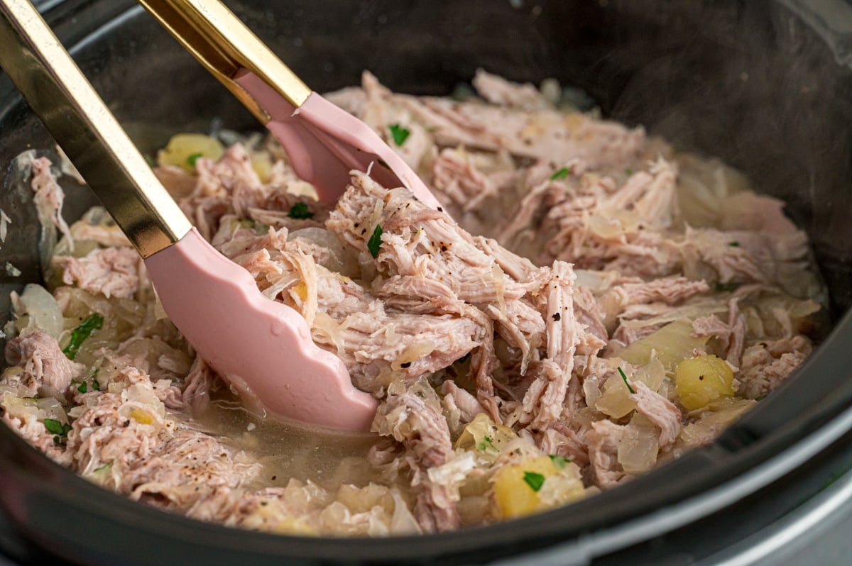 Tongs grabbing some pork and sauerkraut from a slow cooker.