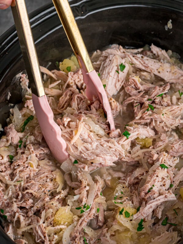 Pork and sauerkraut in a slow cooker with tongs in it.