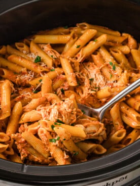 A large spoon scooping chicken pasta rosa from a slow cooker.
