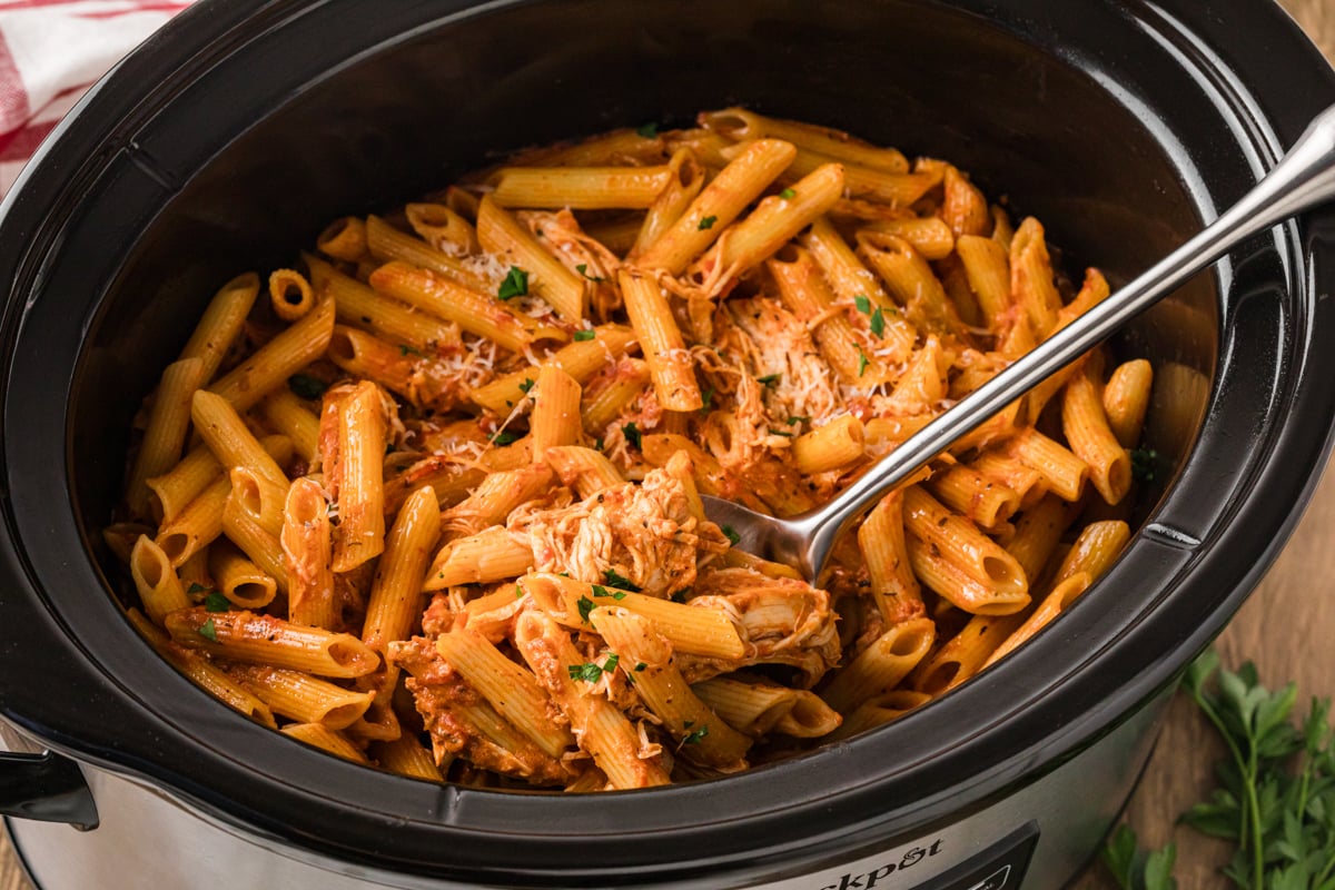 A large spoon scooping chicken pasta rosa from a slow cooker.