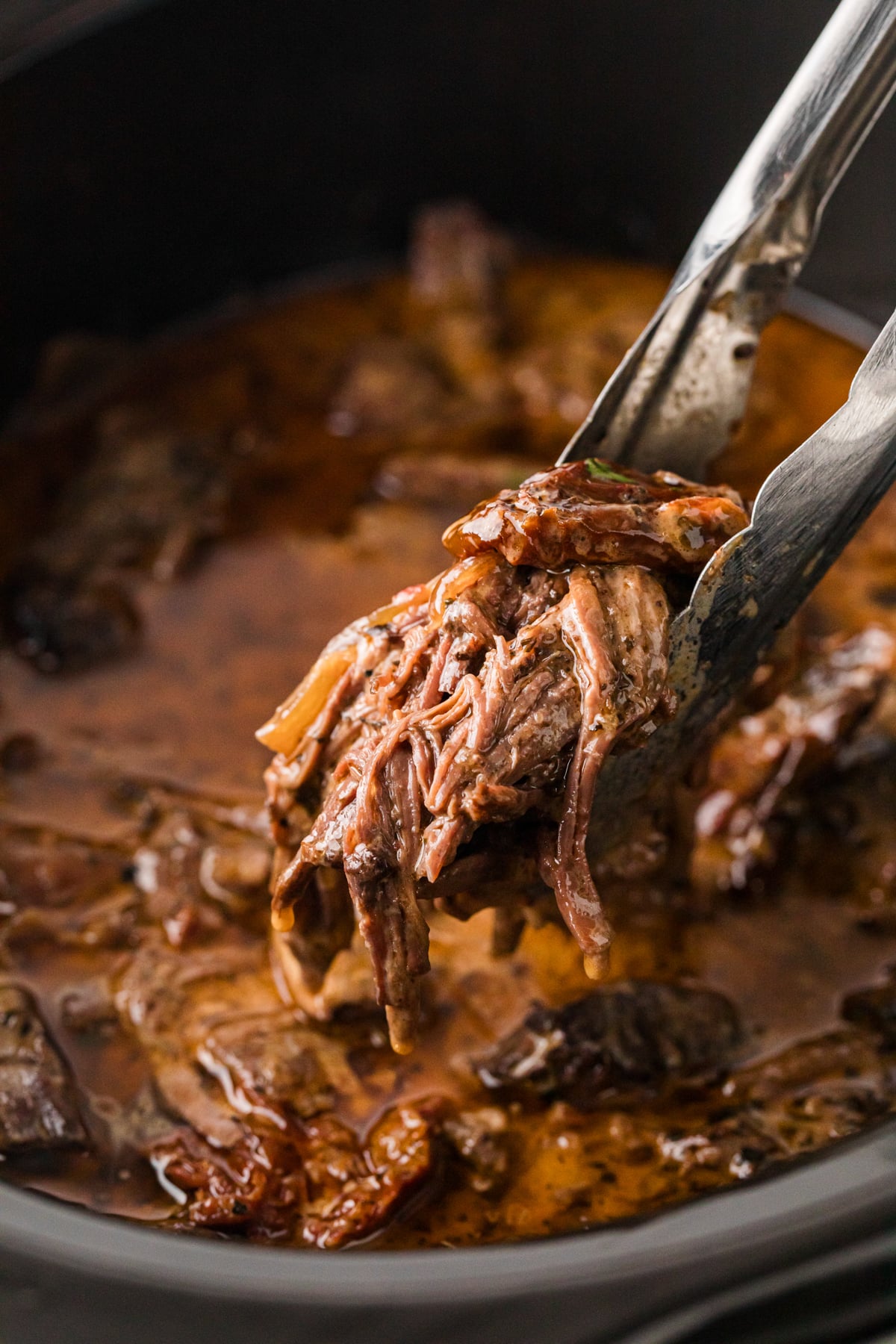 Tongs grabbing shredded pot roast from a crockpot.