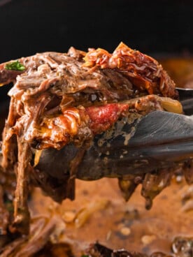 Tongs holding shredded marry me pot roast over a slow cooker.