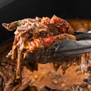 Tongs holding shredded marry me pot roast over a slow cooker.