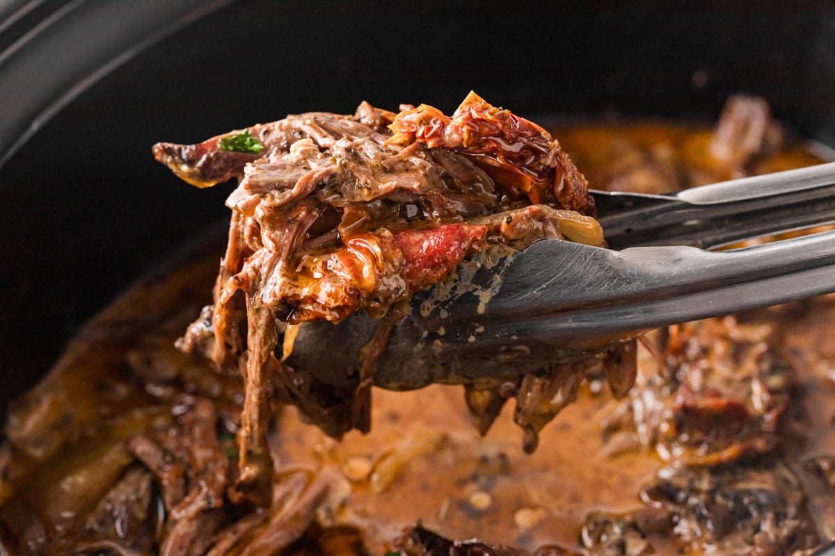Tongs holding shredded marry me pot roast over a slow cooker.