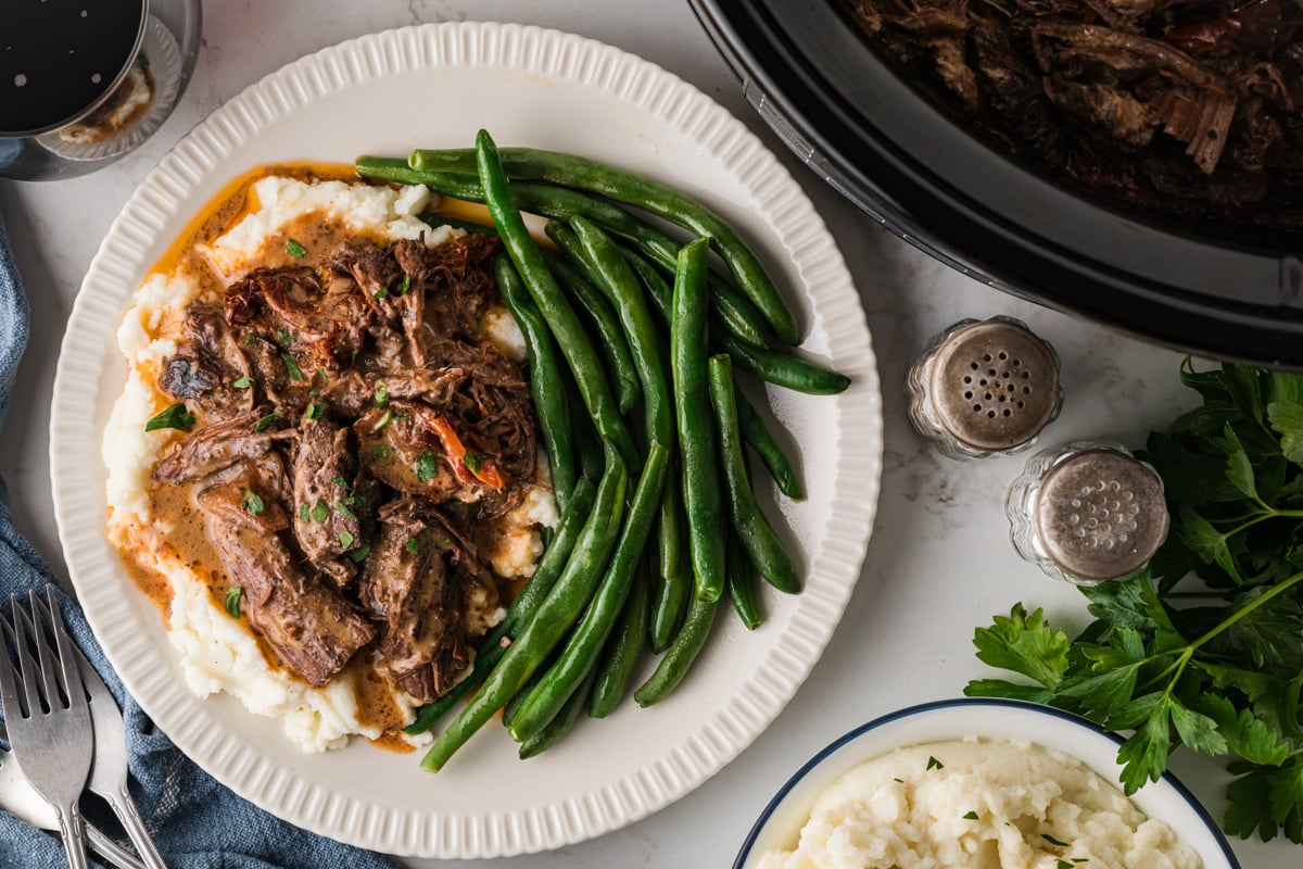 A pot roast dinner with mashed potatoes and green beans on a white plate.