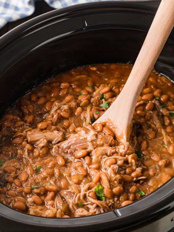 A wooden spoon scooping pork and beans from a slow cooker.