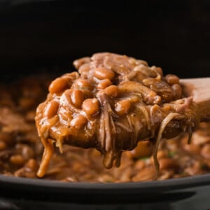 A wooden spoon holding a scoop of pork and beans over a crockpot.