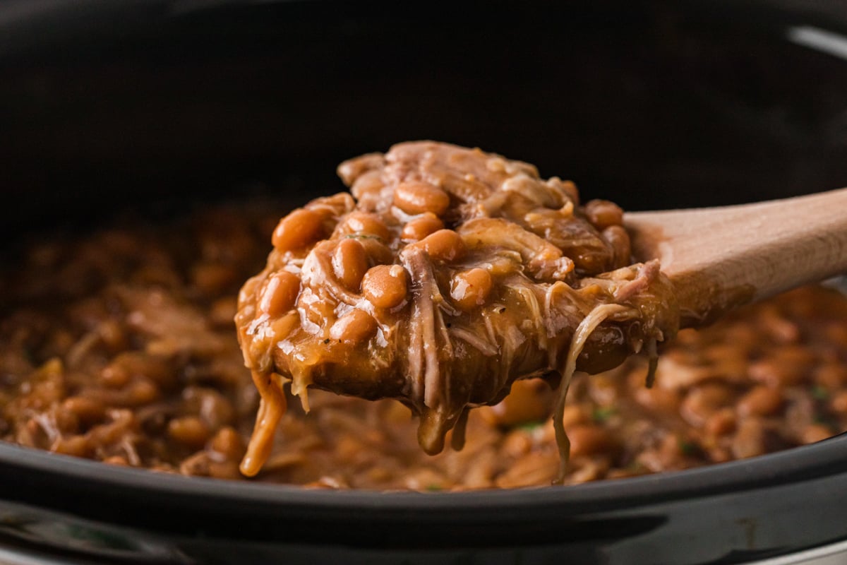 A wooden spoon holding a scoop of pork and beans over a crockpot.