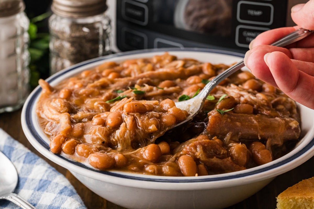 A hand holding a spoonful of baked beans with pork over a bowl.