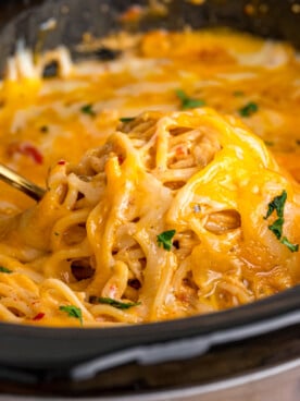 A spoon scooping cheesy spaghetti and chicken from a slow cooker.