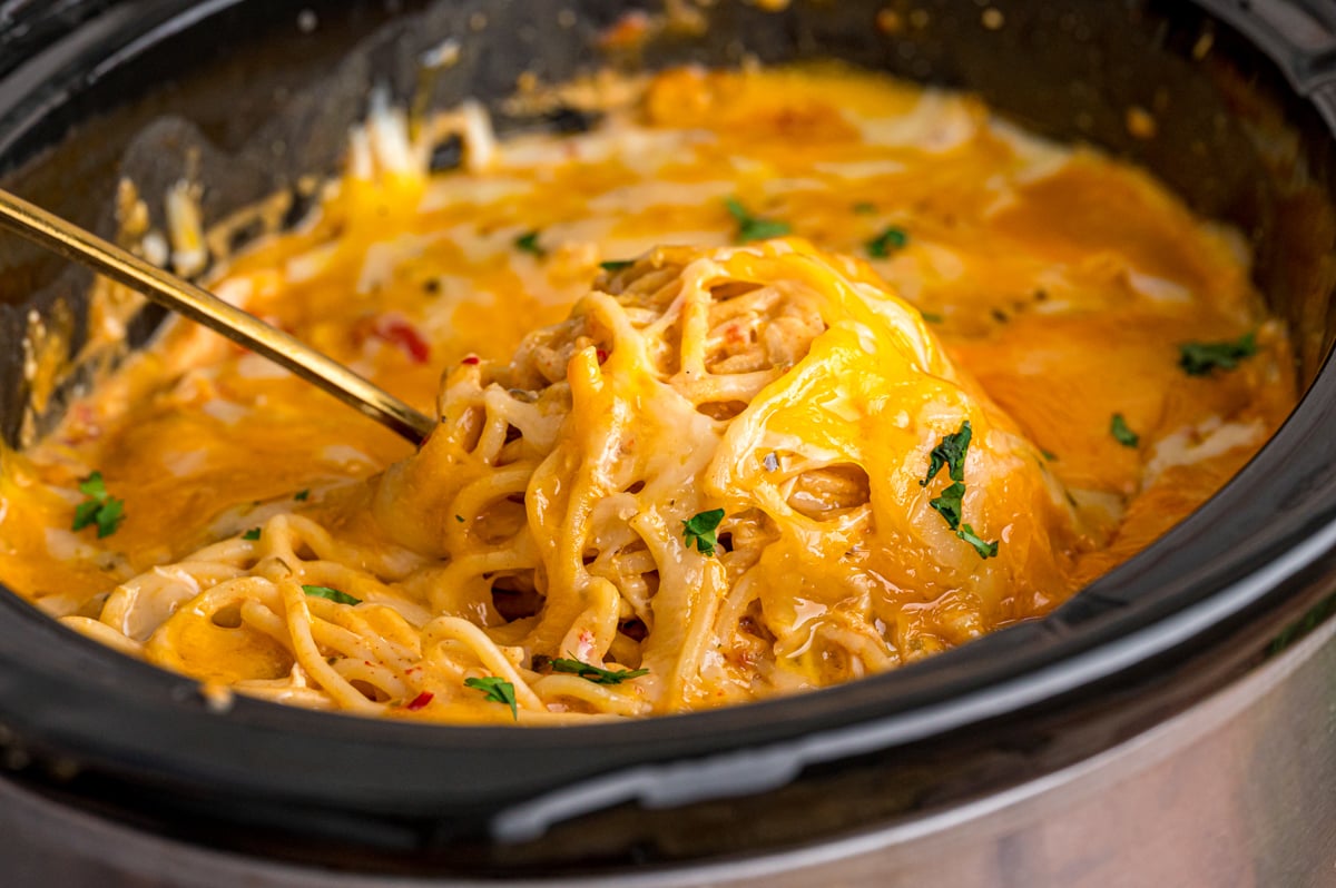 A spoon scooping cheesy spaghetti and chicken from a slow cooker.