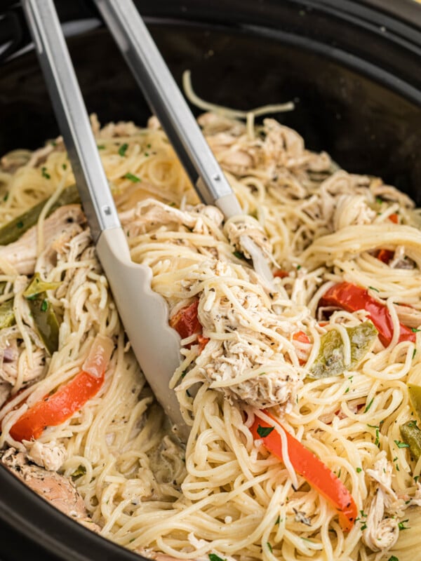 Tongs grabbing chicken and pasta from a slow cooker.