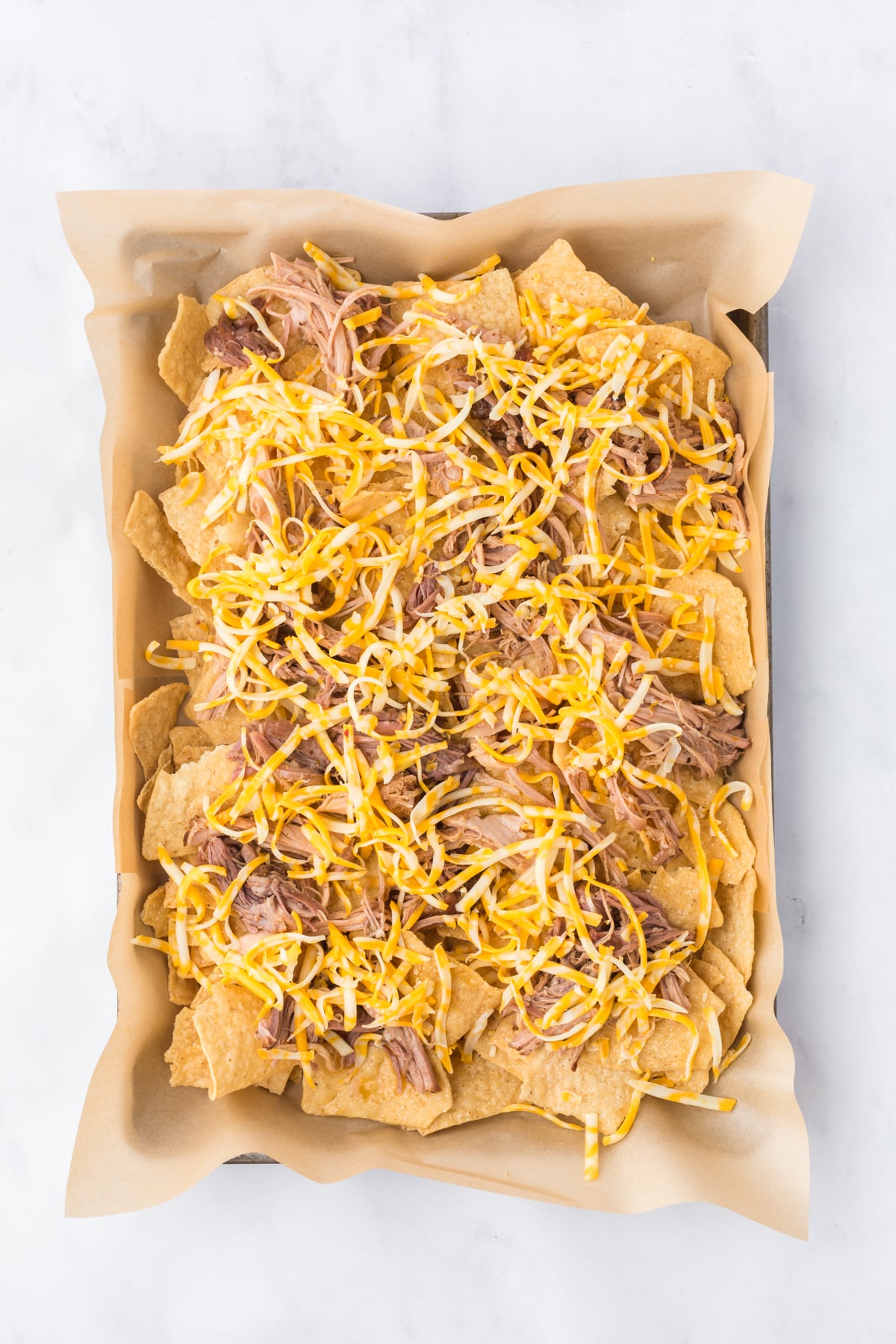 A parchment-lined baking sheet with tortilla chips, pulled pork, and cheese on top.