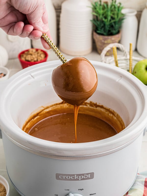 A hand holding an apple over a crockpot as it drips caramel.