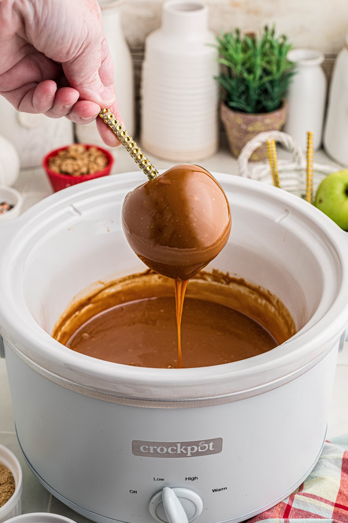 A hand holding an apple over a crockpot as it drips caramel.