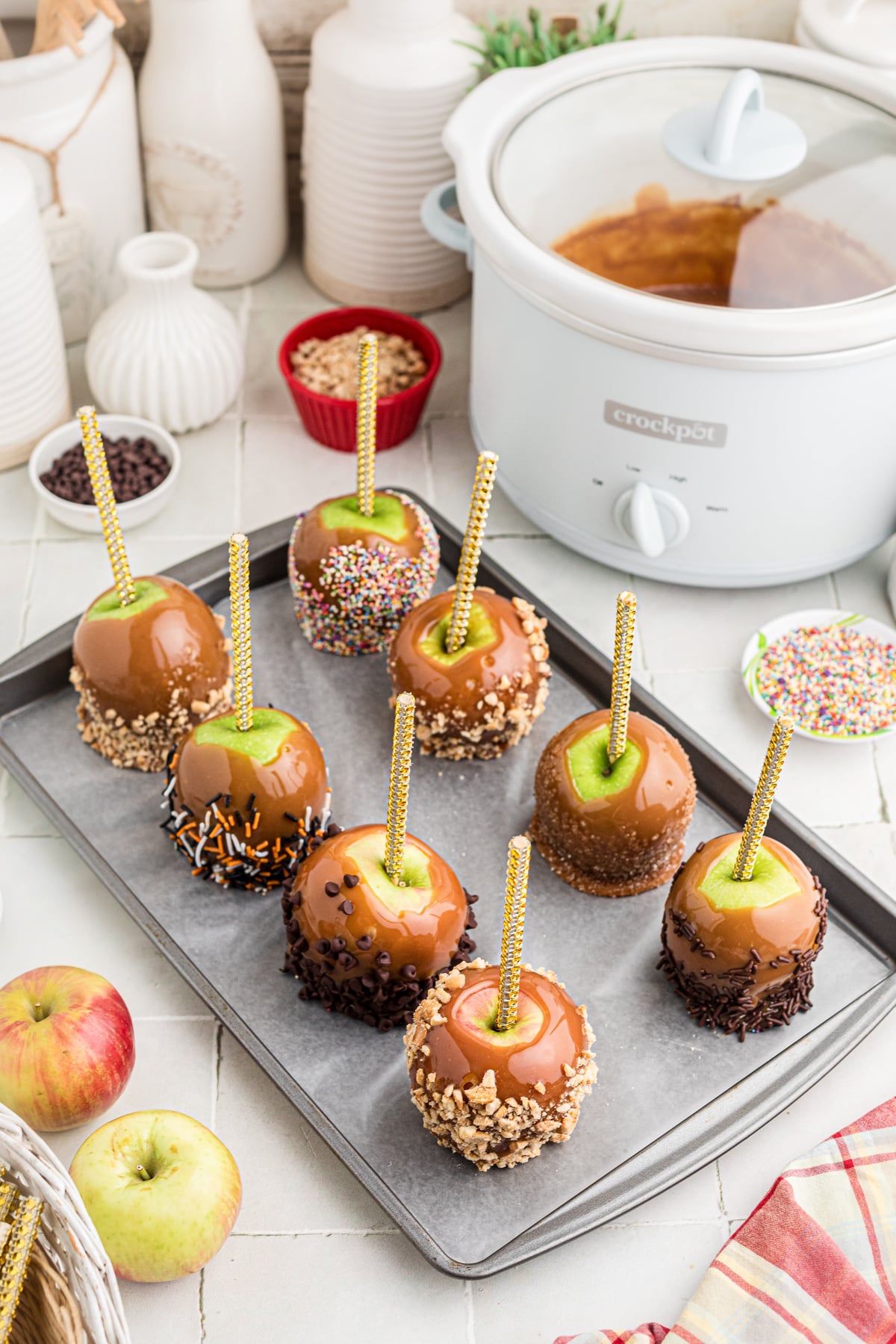 A tray of caramel apples with toppings next to a crockpot of caramel sauce.
